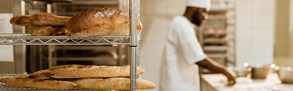 Frisch gebackenes Brot und Baguettes auf einem Regal in einer Bäckerei, während ein Bäcker Teig vorbereitet.