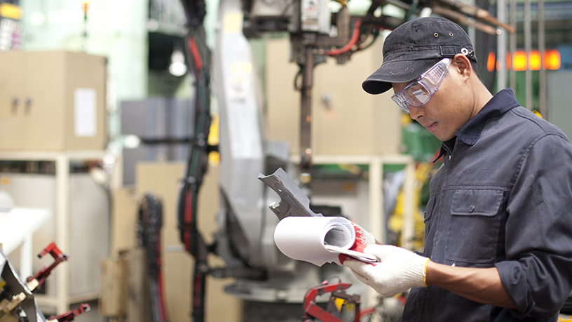 Eine Person prüft technische Unterlagen und hält ein Teil in einer industriellen Umgebung mit Maschinen im Hintergrund.