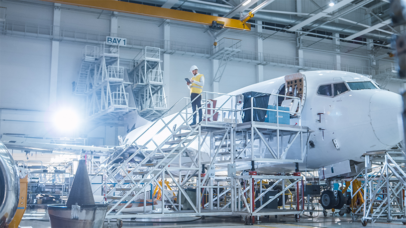 Eine Person steht auf einem Gerüst neben einem Flugzeug und überprüft Dokumente in einer weitläufigen Montagehalle.