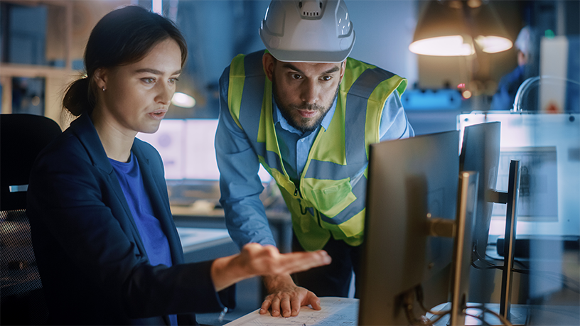 Eine Person zeigt auf Dokumente, während eine andere in Sicherheitskleidung am Computer arbeitet.