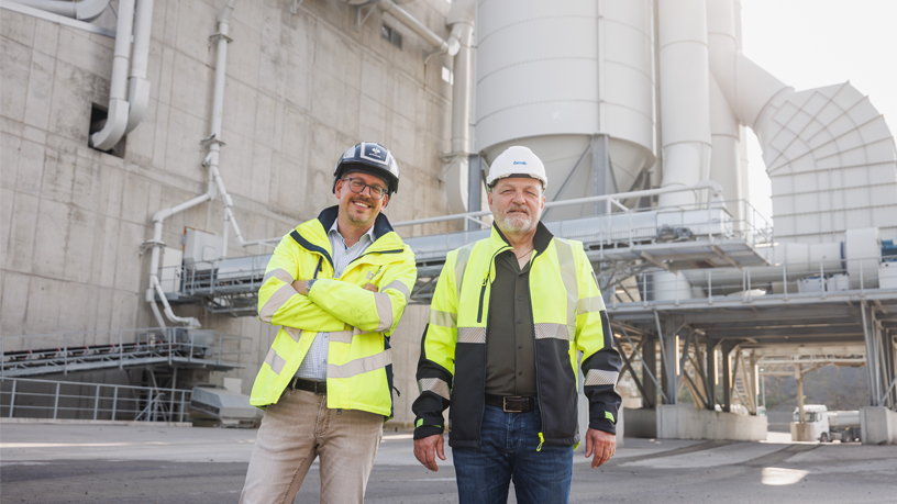 Zwei Personen in Warnwesten und Schutzhelmen stehen vor einer Industrieanlage.