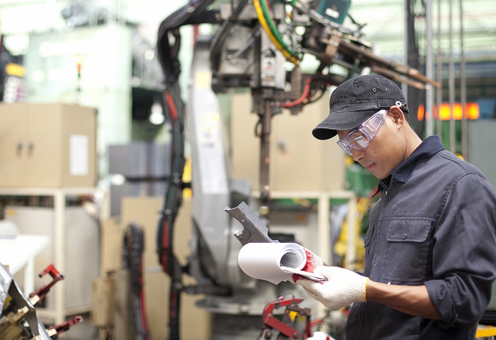 Eine Person überprüft technische Unterlagen und hält ein Bauteil in einer industriellen Umgebung mit Maschinen im Hintergrund.