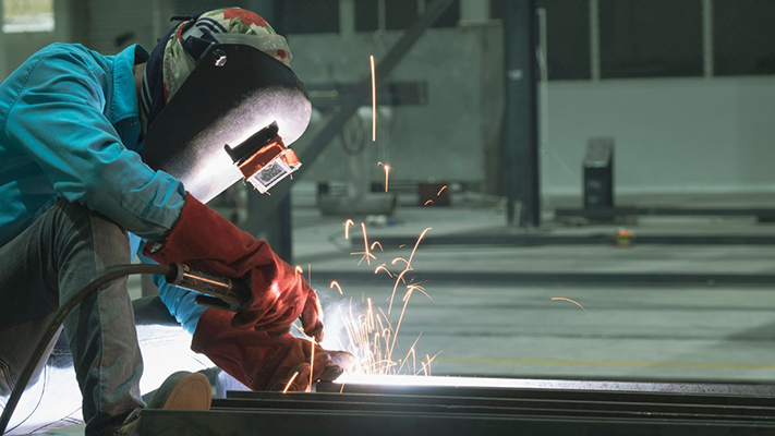 Ein Schweißer in Schutzkleidung und Helm arbeitet an einem Metallstück, Funken fliegen.