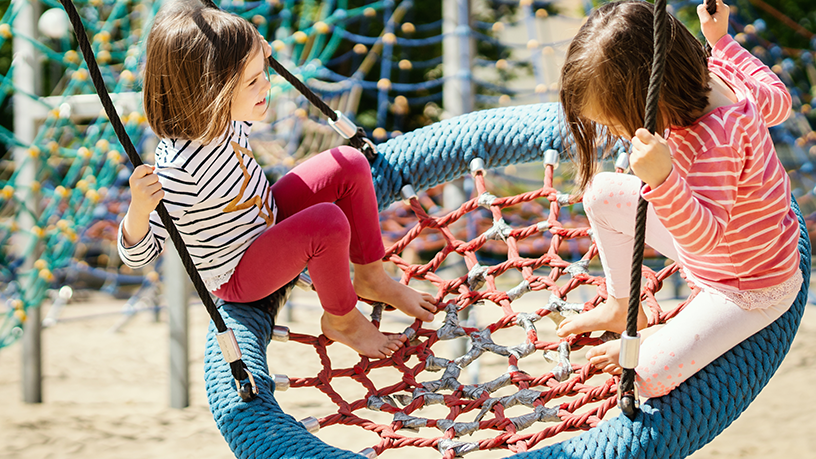 Zwei Kinder spielen auf einer großen Netzschaukel auf einem Spielplatz.