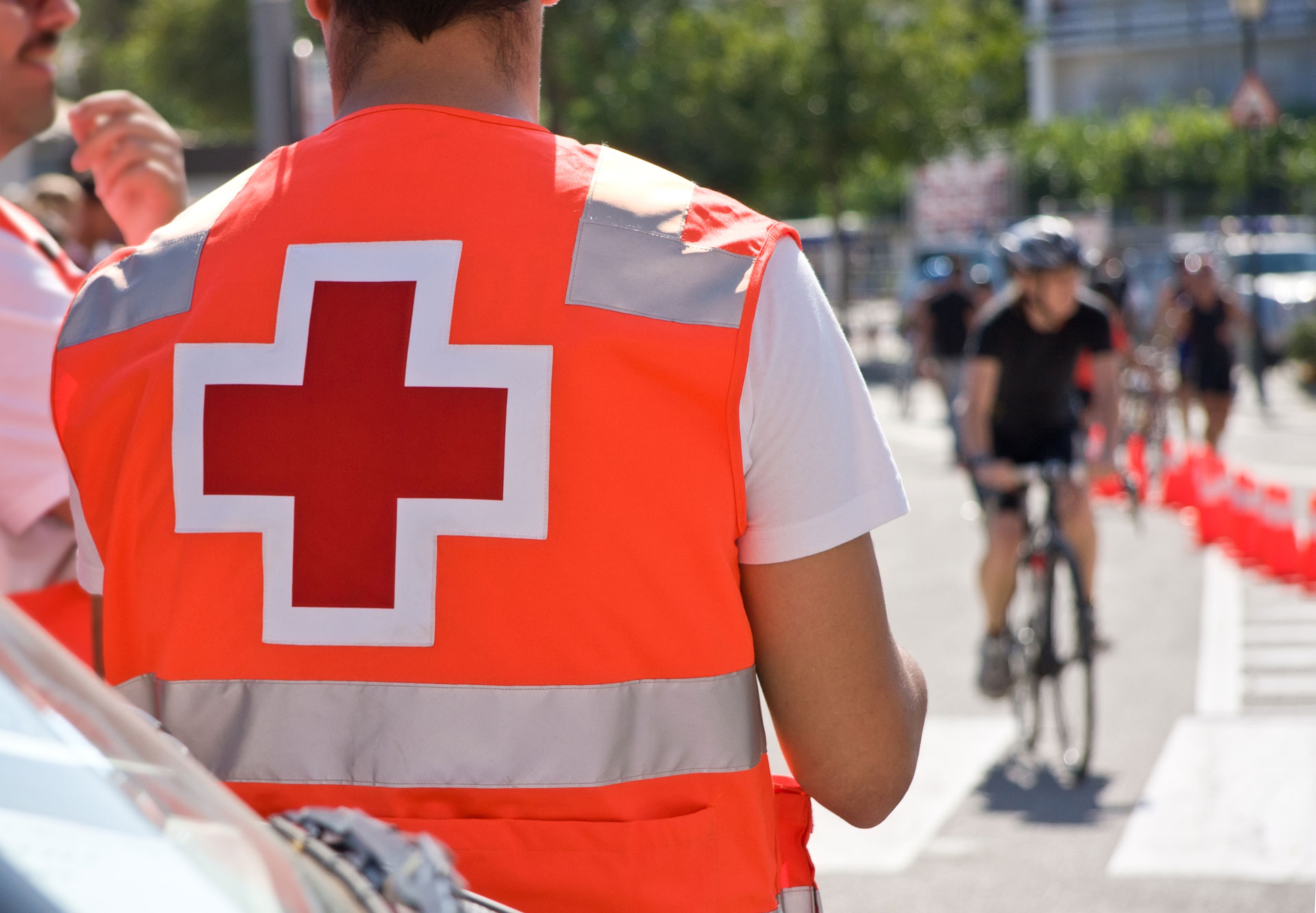 Sanitäter in roter Weste mit Kreuz, im Hintergrund Fahrradfahrer auf Straße