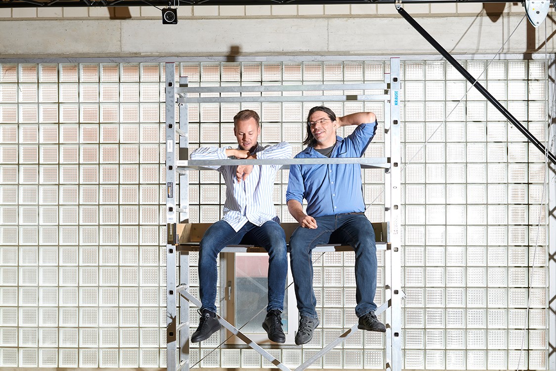 Zwei Personen sitzen auf einem Gerüst vor einer Wand aus Glasbausteinen.