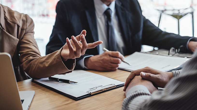 Personen in einem Meeting sitzen am Tisch und diskutieren, mit Klemmbrettern und Dokumenten vor sich.