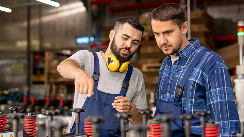 Zwei Arbeiter in einer Fabrik tragen Overalls und Sicherheitsausrüstung, stehen neben Maschinen.