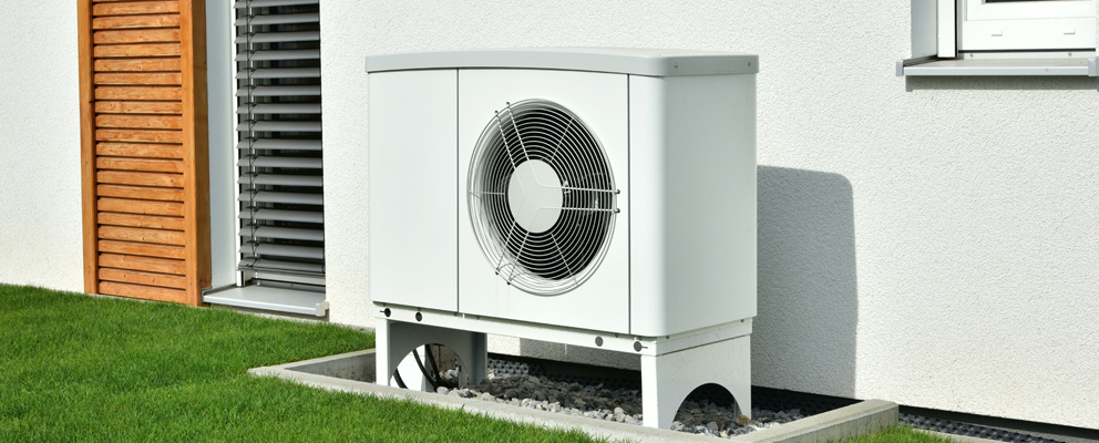 Weiße Wärmepumpe vor Hauswand mit Fenster und Holzverkleidung, auf grünem Rasen.