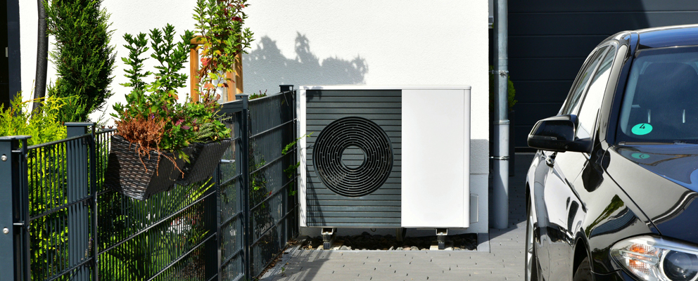 Wärmepumpe neben einem geparkten Auto und Zaun mit Pflanzen vor Hauswand.
