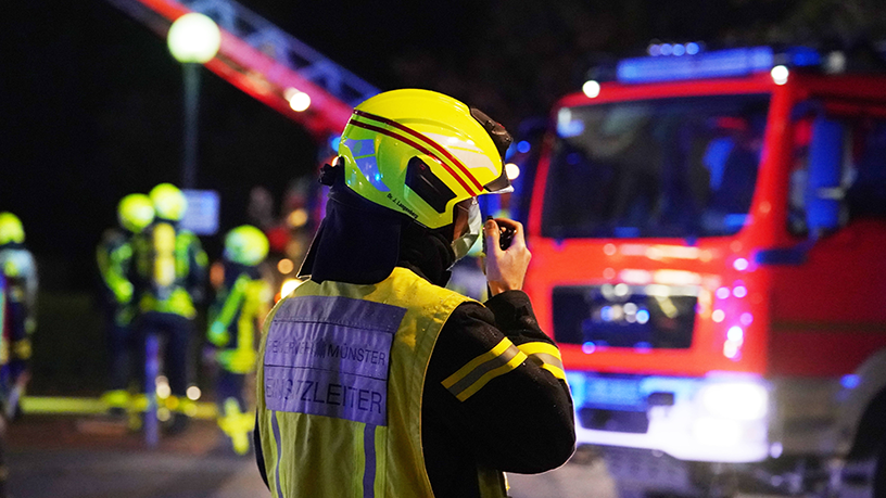 Einsatzleiter der Feuerwehr spricht im Funkgerät, Hintergrund Feuerwehrauto und Feuerwehrleute bei Nacht.