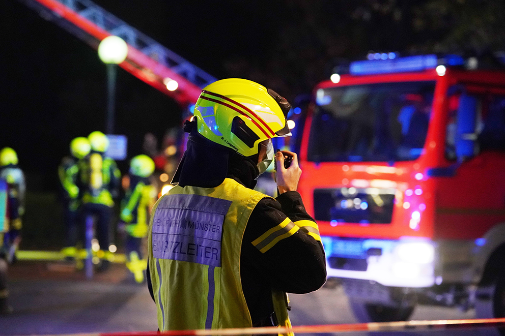 Feuerwehreinsatzleiter kommuniziert per Funkgerät bei einem nächtlichen Einsatz, im Hintergrund Feuerwehrfahrzeug.