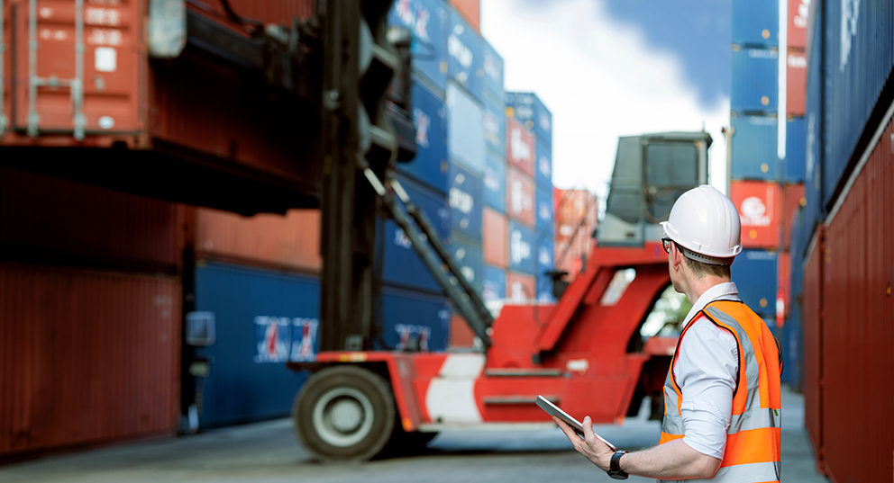 Person in Sicherheitskleidung überwacht Containerverladung mit Tablet in einem Hafen.