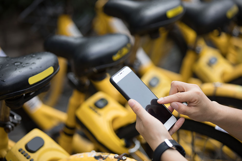 Hände halten ein Smartphone, im Hintergrund gelbe Fahrräder. Sharing Economy