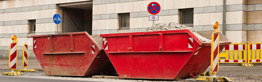Zwei rote Container voller Schutt vor einem Gebäude, umgeben von Baustellenschildern.