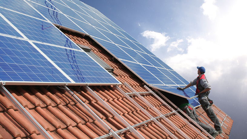 Arbeiter installiert Solarmodule auf einem Ziegeldach unter wolkigem Himmel.