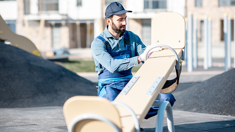Ein Arbeiter in blauer Arbeitskleidung und Mütze überprüft eine Wippe auf einem Spielplatz.