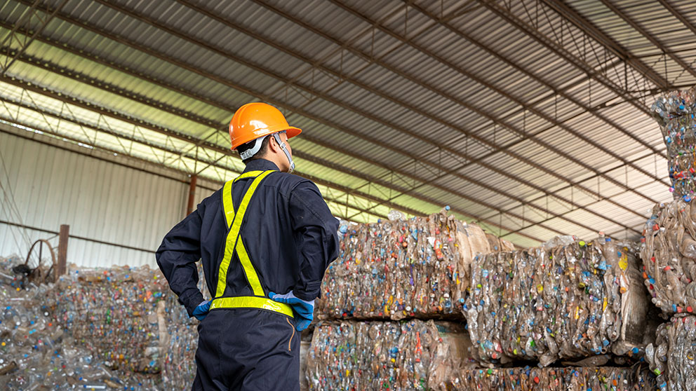 Ein Arbeiter in Schutzkleidung steht in einer Recyclinganlage vor gestapelten gepressten Plastikflaschen.