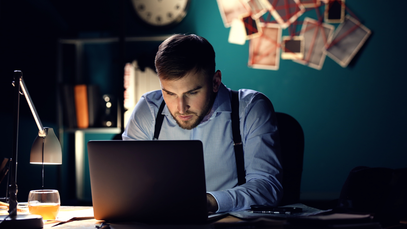 Eine Person sitzt nachts an einem Schreibtisch und arbeitet am Laptop, umgeben von Dokumenten und alkoholischem Getränk.