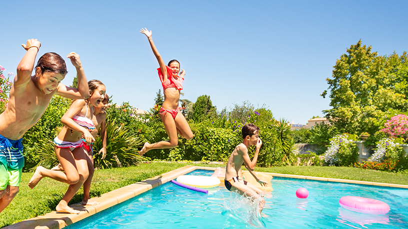 Kinder springen fröhlich in einen Swimmingpool, umgeben von grünen Pflanzen und bunten Schwimmreifen.
