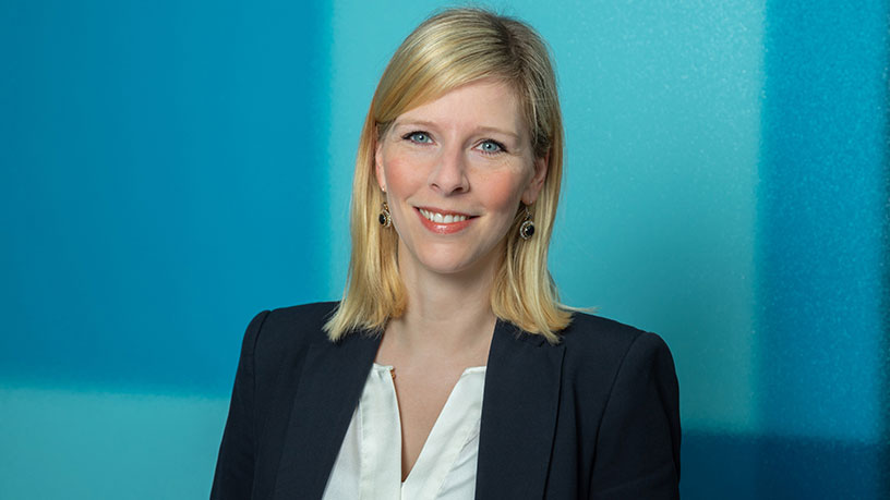 Catharina Fröhling in einem schwarzen Blazer und weißer Bluse vor einem blauen Hintergrund.