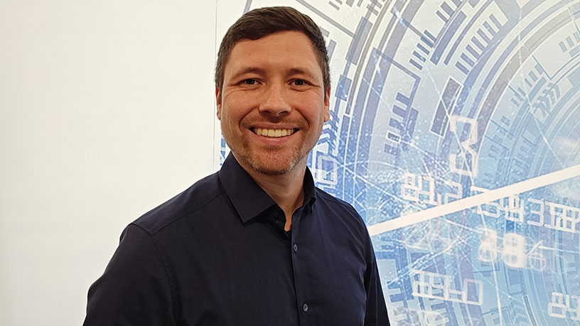 Dr. Tim Brückmann steht vor einem digitalen, technologisch anmutenden Hintergrund in einem dunkelblauen Hemd.