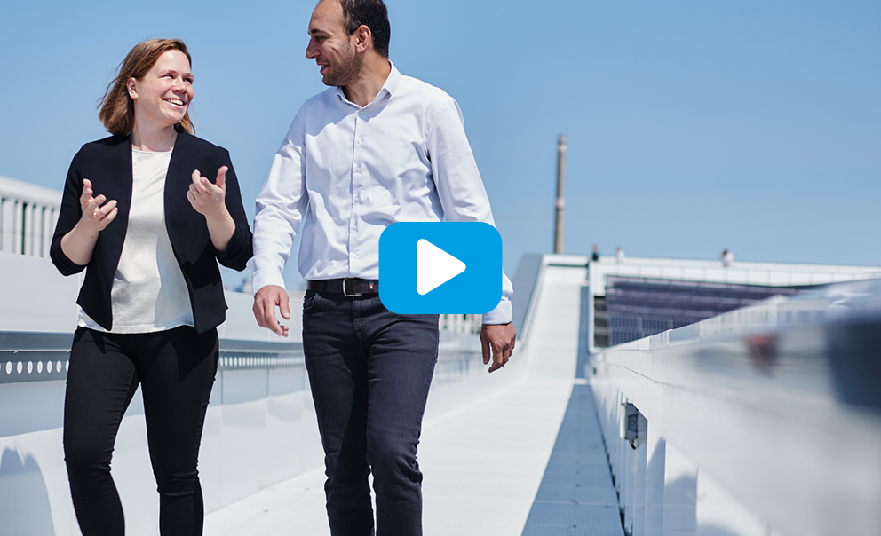 Zwei Personen gehen und sprechen auf einem modernen Gehweg; Play-Button zeigt Video an.