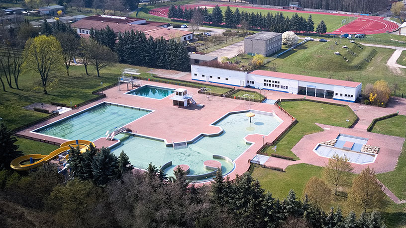 Luftaufnahme eines Freibads mit mehreren Schwimmbecken, einer Wasserrutsche und umliegender Grünfläche.