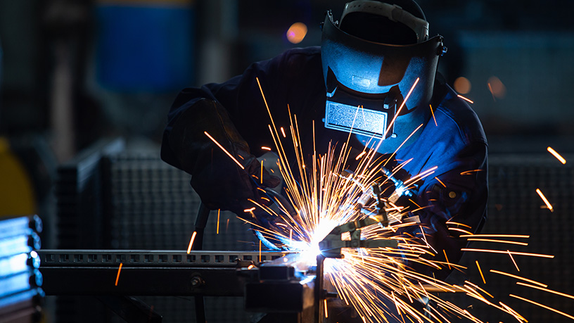 Schweißer mit Schutzhelm und -kleidung arbeitet an einem Metallstück, umgeben von hellen Funken.
