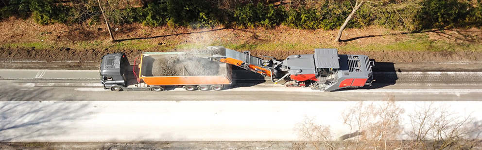 Luftaufnahme einer Straßenbaumaschine, die Material in einen orangefarbenen Lkw kippt.