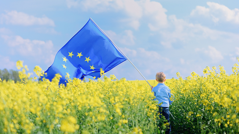 Ein Kind steht in einem blühenden Rapsfeld und hält eine Europaflagge.