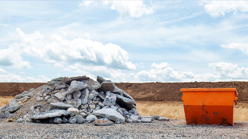 Steinhaufen neben einem orangefarbenen Container auf einer Baustelle unter einem bewölkten Himmel.
