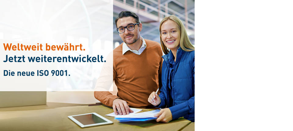 Personen arbeiten an einem Tisch mit Dokumenten und Tablet in einer Fabrikhalle, Überlagernd Text zur neuen ISO 9001-Norm.