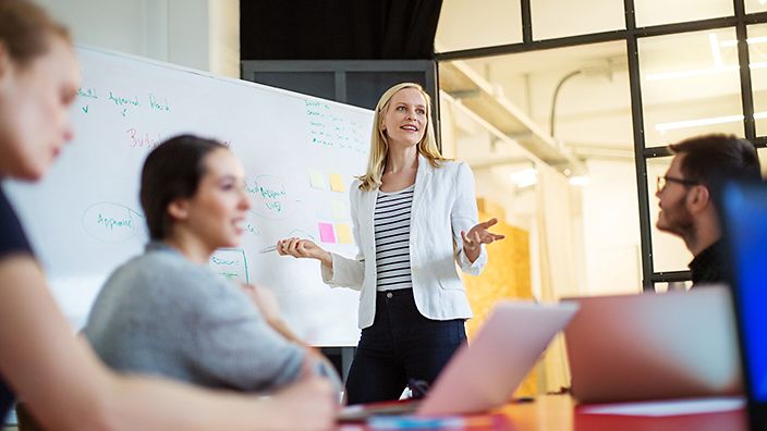 Person hält Präsentation vor Gruppe, mit Fokus auf Management und Qualitätsstrategien.