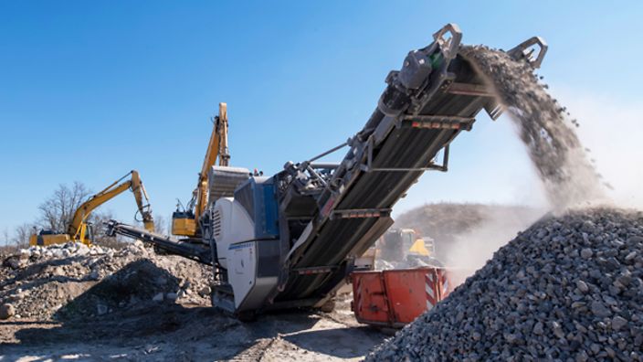Baufahrzeuge auf Baustelle, Gestein wird durch Förderband bewegt.