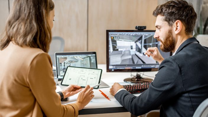 Zwei Personen arbeiten an Computern mit technischen Zeichnungen.